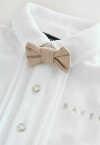 Valkoinen teksturoitu paita, jossa on käännetty kaulus, beigen värinen rusetti ja hopeiset napit, joissa on "BAKER"-brändäys.