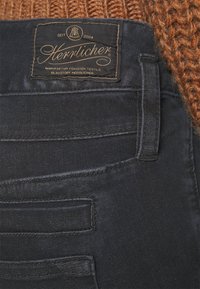 Närbild av en ficka på mörka jeans med en svart rektangulär Herrlicher-märke och en senapsgul stickad tröjkant ovanför.