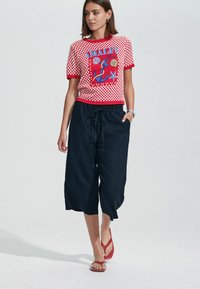 Raudona ir balta languota marškinė su ankerio grafika, derinama su tamsiai mėlynomis trumpomis kelnėmis ir raudonais šlepetėmis. Kasdienių vasaros drabužių derinys.