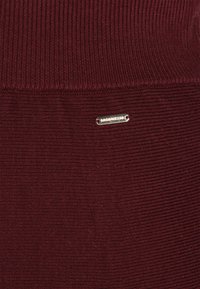 Burgundy ribbed triko-kangas, jossa on hopeinen metallilogo-merkki ommeltuna alapuolelle. Sujuva pinta ja tasainen kuvio.