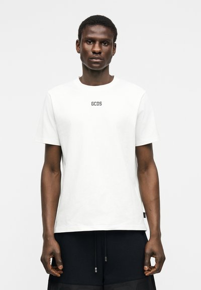 Νεαρός άνδρας με σκουρόχρωμο δέρμα, ντυμένος με λευκό t-shirt GCDS και μαύρο παντελόνι, στέκεται μπροστά σε απλό ανοιχτό γκρι φόντο.