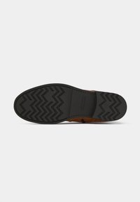 Zapato de cuero marrón con una superficie texturizada, que presenta una suela de goma negra. La suela tiene un patrón en zigzag para mejorar la tracción.