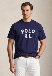 Άνδρας που φοράει μπλε σκούρο t-shirt "POLO R.L.", άσπρο παντελόνι, καφέ ζώνη και μπλε ρολόι χειρός, στέκεται με το ένα χέρι στην τσέπη.
