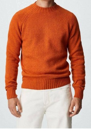 Mann trägt einen enganliegenden orangen Strickpullover mit gerippten Bündchen und Saum, kombiniert mit weißen Hosen, steht vor einem schlichten Hintergrund.