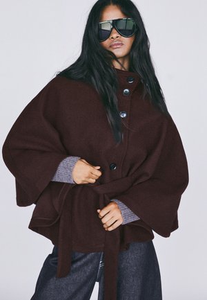 Femme aux longs cheveux foncés portant une cape marron oversize boutonnée avec ceinture, manches grises, jean foncé et grandes lunettes de soleil noires.