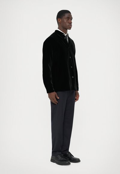 Black velvet button-up jacket with a curved collar, paired with navy trousers and black lace-up shoes. Smooth texture, minimal design.