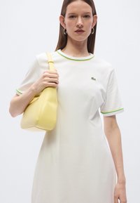 Λευκό βαμβακερό φόρεμα με πράσινη λεπτομέρεια, με λογότυπο Lacoste. Κίτρινη τσάντα ώμου με απαλή υφή, κυρτό σχήμα και μινιμαλιστικό σχεδιασμό.