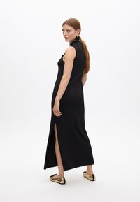 Vestido maxi negro sin mangas con abertura lateral, diseño ajustado, textura suave, con un cuello alto y acentos brillantes. Se lleva con sandalias planas doradas.