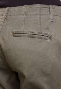 Pantalons vert olive à la texture lisse, avec une poche arrière à rabat et un détail de couture.