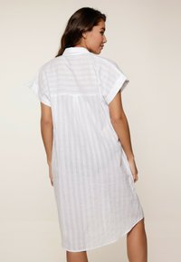 Abito camicia in lino bianco con maniche corte arrotolate, caratterizzato da righe orizzontali e dettagli a occhiello, lungo fino al ginocchio con una leggera fessura laterale.