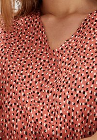 Halenka s výstřihem do V na korálovém pozadí, zdobená rozptýleným vzorem černých a bílých teček. Vyrobena z měkkého, lehkého materiálu.