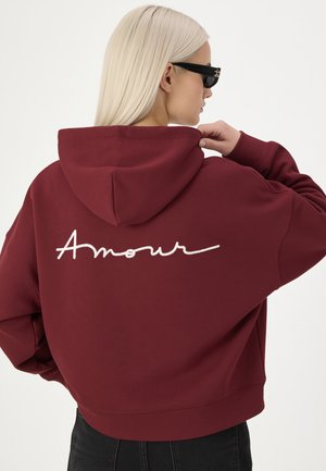Vrouw met lang blond haar, zwarte zonnebril en een bordeauxrode hoodie met het woord "Amour" in witte letters op de achterkant.