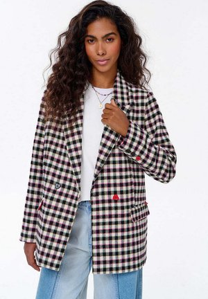 Žena s dlouhými kudrnatými vlasy má na sobě černobíle růžový kostkovaný blejzr přes bílé tričko a světle modré džíny, drží si klopu blejzru.