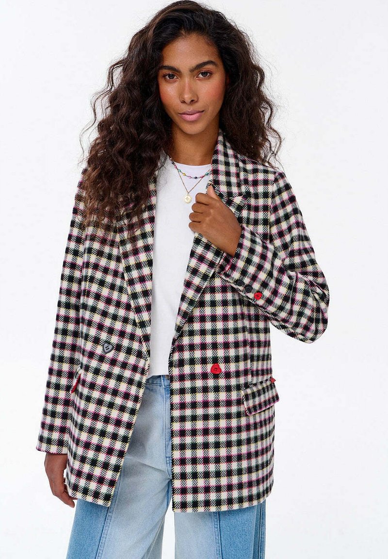Žena s dlouhými kudrnatými vlasy má na sobě černobíle růžový kostkovaný blejzr přes bílé tričko a světle modré džíny, drží si klopu blejzru.