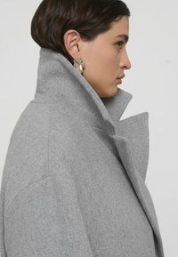 Cappotto grigio in misto lana con collo alto e design strutturato. Presenta rever angolari e una finitura morbida e testurizzata.