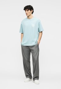 Νέος άνδρας με καστανά σγουρά μαλλιά που φοράει ένα ανοιχτό μπλε φαρδύ μπλουζάκι, γκρι παντελόνι με φαρδιά μπάσκα και λευκά αθλητικά παπούτσια, στέκεται με τα χέρια στις τσέπες.