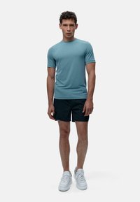 Kortärmad turkos träningströja med rund halsringning, tillsammans med svarta shorts och vita sneakers. Slät tyg med subtila design detaljer.