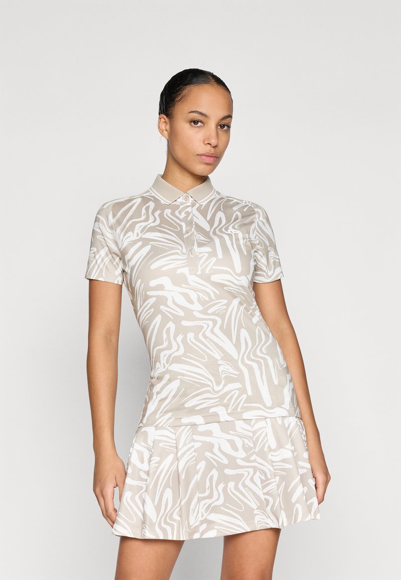 Přiléhavé béžové polo šaty s bílými abstraktními vzory, krátké rukávy a skládací sukně. Mají klasický límeček a knoflíkovou légou.