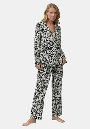 Kobieta stoi boso, ubrana w czarno-białą, w kropki, koszulę z długim rękawem zapinaną na guziki oraz dopasowane szerokie spodnie.