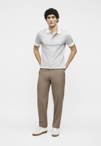 Νέος άνδρας στέκεται με τα χέρια στις τσέπες, φοράει λευκό πόλο πουκάμισο, καφέ παντελόνι και λευκά αθλητικά παπούτσια σε απλό φόντο.