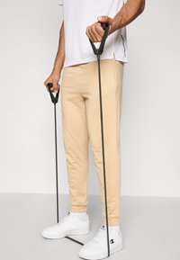 Bézs sportnadrág és fehér ing, fekete ellenállási szalagokat tartva. Fehér sportcipők világosszürke részletekkel, a szalagokon texturált fogantyúk.