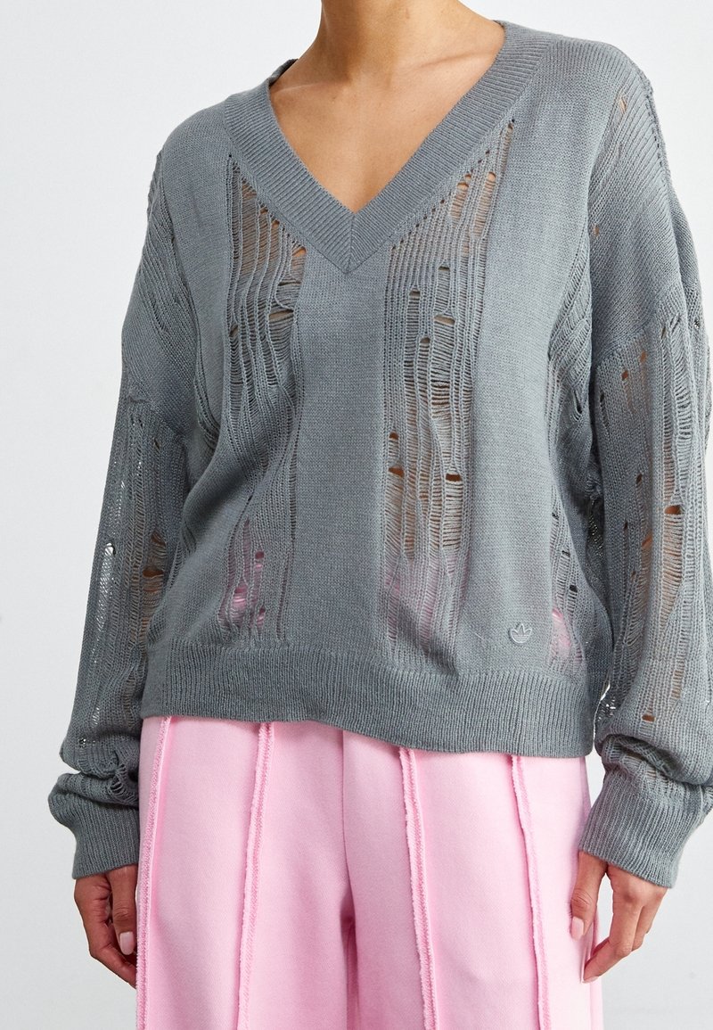 Grå stickad tröja med V-ringning och slitna detaljer, med ribbat avslut och mudd. Diskret logotyp längst ner fram.