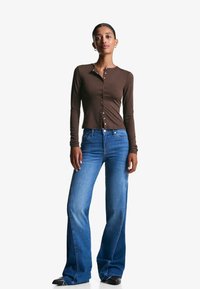 Bruine langemouwen knooptop met een getailleerd ontwerp, gecombineerd met blauwe uitlopende jeans en zwarte schoenen, tegen een effen witte achtergrond.