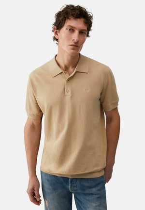 Uomo con capelli ricci che indossa una polo beige a maniche corte e jeans blu, in piedi contro uno sfondo chiaro e uniforme.