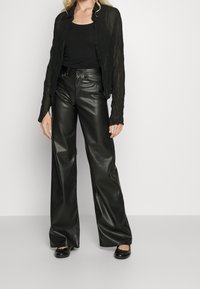 Musta läpinäkyvä, ryppyinen takki mustan toppipaidan päällä, yhdistettynä korkeavyötäröisiin, leveälahkeisiin keinonahkahousuihin ja mustiin ballerinatossuihin.