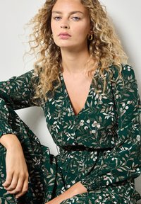 Groene jurk met een bladpatroon, voorzien van een diepe V-hals, knopen aan de voorkant en lange mouwen met elastische manchetten. Zachte textuur.