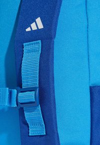 Modrá část batohu s texturovanou tkaninou, modrým zapínáním a bílým logem. Hladký povrch s vyztuženými švy.