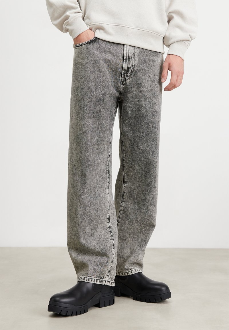 Jeans de mezclilla gris claro con un ajuste relajado, que presentan un acabado deslavado, costuras prominentes y bolsillos. Combinados con zuecos negros gruesos.