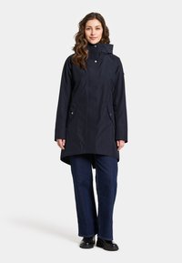 Marin vattentät jacka med hög krage, två framfickor och en något längre bakstycke. Slät textur och funktionell design.