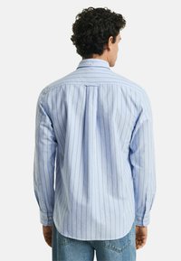 Camicia a righe blu chiaro in cotone, con maniche lunghe, colletto con bottoni e un pieghettato sul retro per facilitare i movimenti.