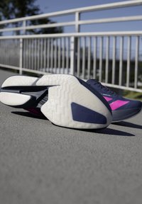 Chaussures de course avec semelles blanches texturées et accents violet-rose posées sur le trottoir près d'une clôture en métal sous un ciel dégagé.