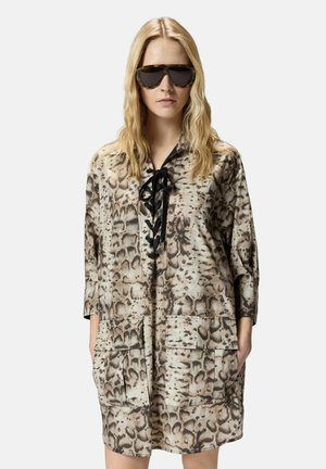 Blondine draagt een oversized jurk met slangenprint, voorzakken en zwarte veters, gecombineerd met grote zonnebrillen in schildpadmotief.
