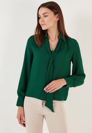 Blusa verde scuro con colletto e dettagli a cravatta, realizzata in tessuto morbido. Caratterizzata da maniche lunghe e polsini con bottoni.