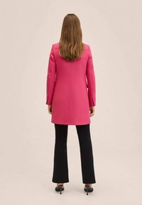 Abrigo rosa con un diseño ajustado, mangas largas y corte recto, combinado con pantalones negros acampanados y tacones. Acabado de tela suave.