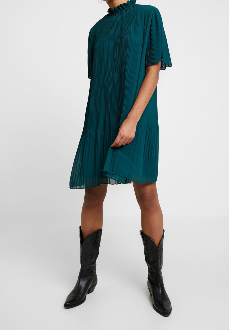 Teal-harmaari mekko, jossa on korkea röyhelökaulus ja lyhyet hihat, yhdistettynä mustiin cowboy-kenkäihin, joissa on terävä kärki ja ompeluyksityiskohtia.