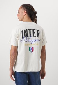 Kremfarget t-skjorte med svart tekst "INTER" og "Nerazzurri." Har et flerfarget emblem med "20." Korte ermer, avslappet passform.