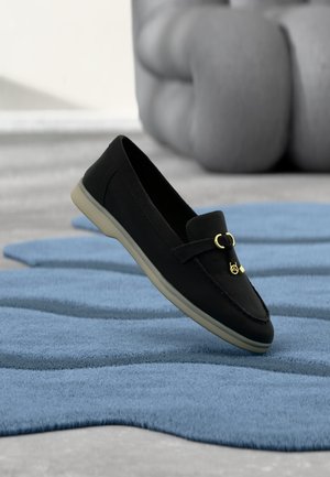Mocasín negro sin cordones con detalle de hebilla dorada sobre una alfombra texturizada azul, cojín desenfocado al fondo sobre suelo gris.