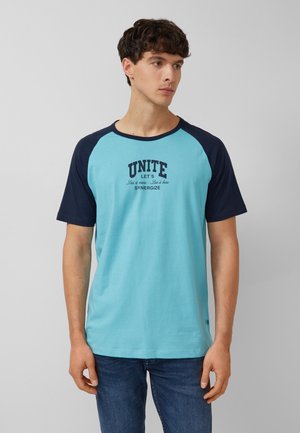 Junger Mann trägt ein hellblaues und marineblaues Raglan-T-Shirt mit dem Schriftzug "UNITE LET'S SYNERGIZE" und steht vor einem einfarbigen grauen Hintergrund.