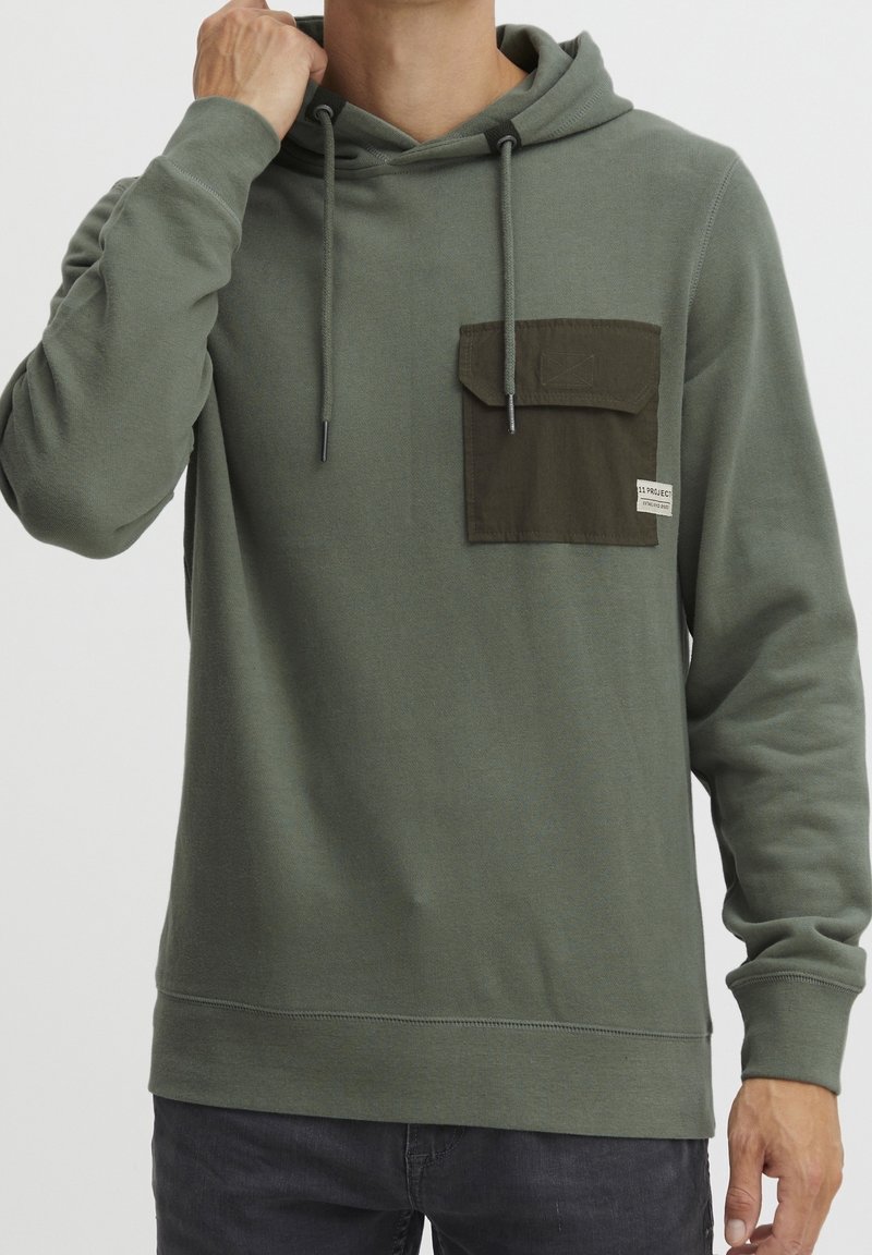 Homme ajustant un sweat à capuche vert olive avec une poche poitrine vert foncé et des cordons, portant un jean foncé devant un fond uni.