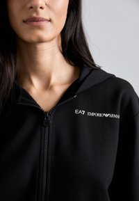 Zwarte rits hoodie gemaakt van zachte stof, heeft een logo op de borst en beschikt over een gladde rits en capuchon.