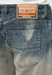 Μπλε τζιν παντελόνι με ξεθωριασμένη υφή, με καφέ δερμάτινη ετικέτα και πίσω τσέπη, που παρουσιάζει υφαντό μοτίβο και φθαρμένες λεπτομέρειες.