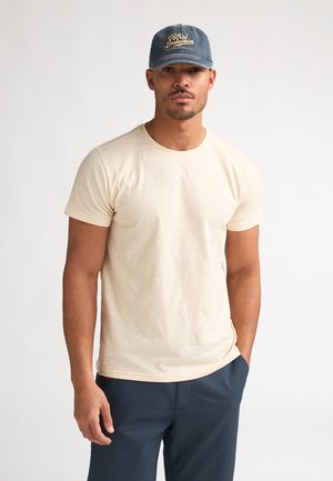 Beige T-shirt met ronde hals en korte mouwen, gecombineerd met marineblauwe broek. Een blauwe pet met beige geborduurde tekst aan de voorkant.