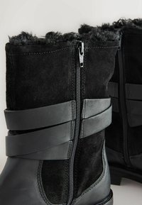 Stivali neri alla caviglia con esterno in suede, dotati di colletto in pelo sintetico, chiusura con zip e cinturini in pelle sulla parte superiore per un tocco di dettaglio.