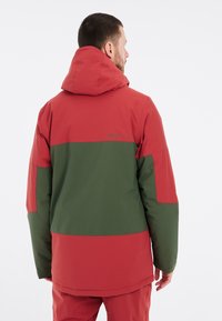 Rot und grün farblich abgesetzte Jacke mit Kapuze, aus einem glatten, wasserdichten Material und mit lockerem Schnitt. Branding auf der Rückseite sichtbar.