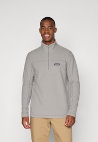 Suéter de forro polar gris claro con un cuello alto, cremallera en la parte frontal hasta la mitad, y un pequeño logo azul en el pecho, que presenta mangas largas y una textura suave.
