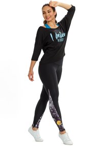 Fekete oversized ing kék felirattal és háromnegyedes ujjakkal, fekete leggings-szel párosítva, amelyek színes virágmintásak az aljukon. Fehér sportcipő.